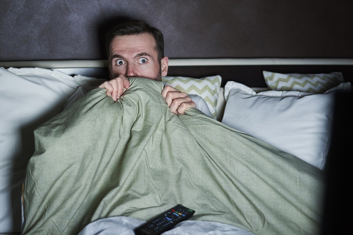 Foto de homem deitado em uma cama, com travesseiros. Ele cobre parte do rosto com um edredom e est&aacute; com a cara de assustado - Metr&oacute;poles