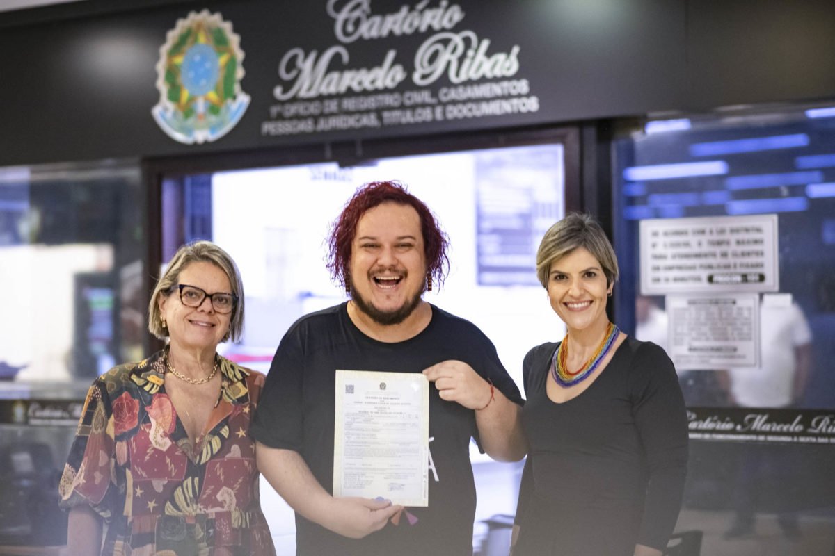 Foto colorida de homem branco de cabelo pintado de ruivo em frente ao cartório segurando a certidão ao lado de duas mulheres: uma mais velha de roupa estampada e outra de roupa preta