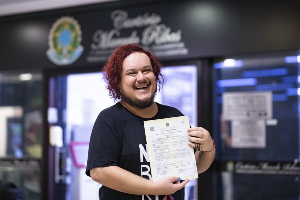 Foto colorida de homem branco de cabelo pintado de ruivo em frente ao cartório segurando a certodão