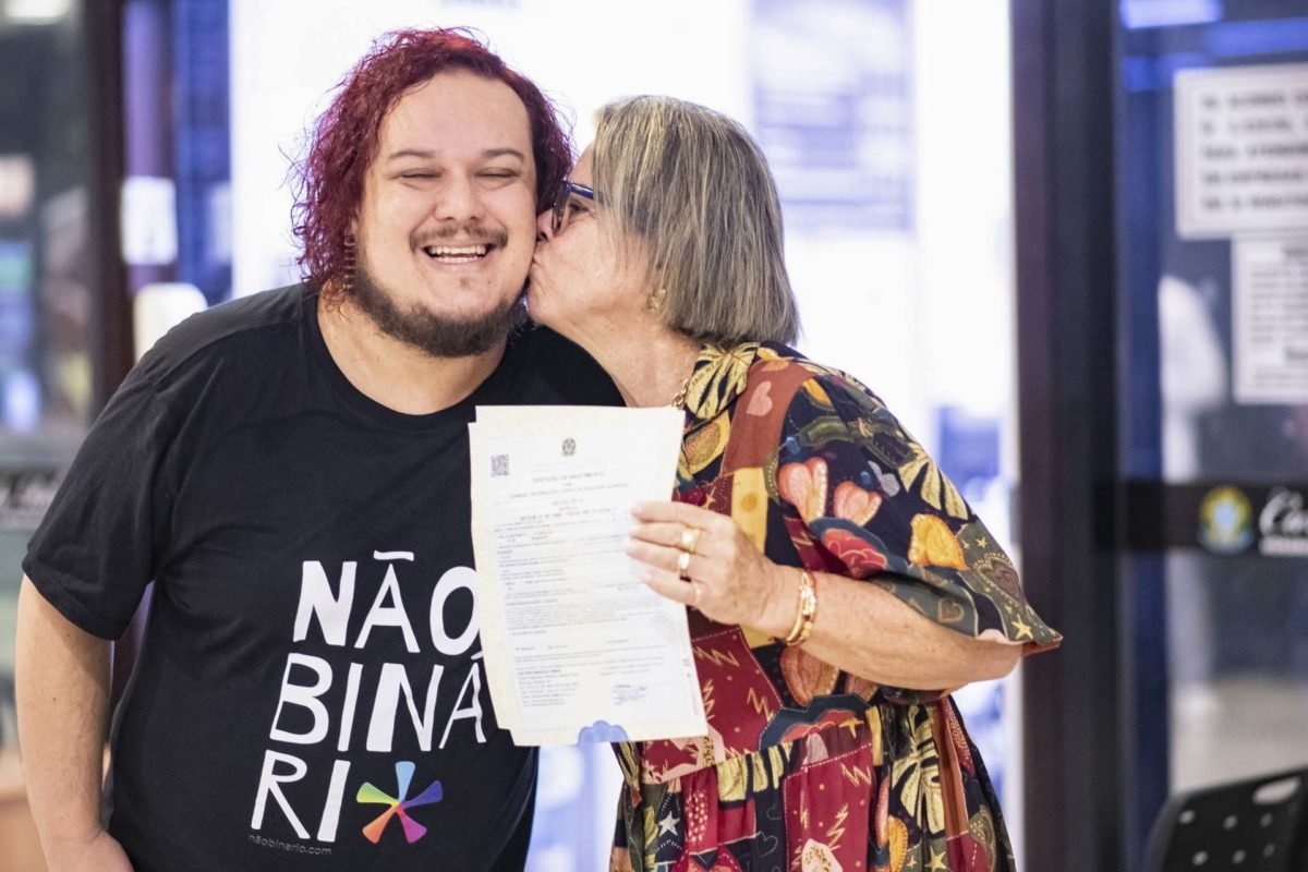 Foto colorida de homem branco de cabelo pintado de ruivo recebendo um beijo na bochecha da mãe, uma mulher branca com roupa estampada segurando certidão