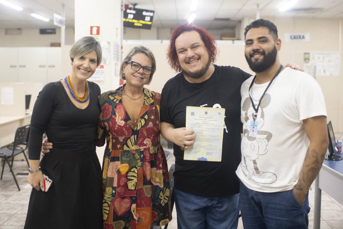 Homem branco de cabelo pintado de ruivo olhando para documento ao lado de mulher branca com cabelo curto e roupa perta e ao lado de mulher mais velha segurando a certidão. Do lado do homem de cabelo vermelho há um homem pardo com blusa branca e barba feita