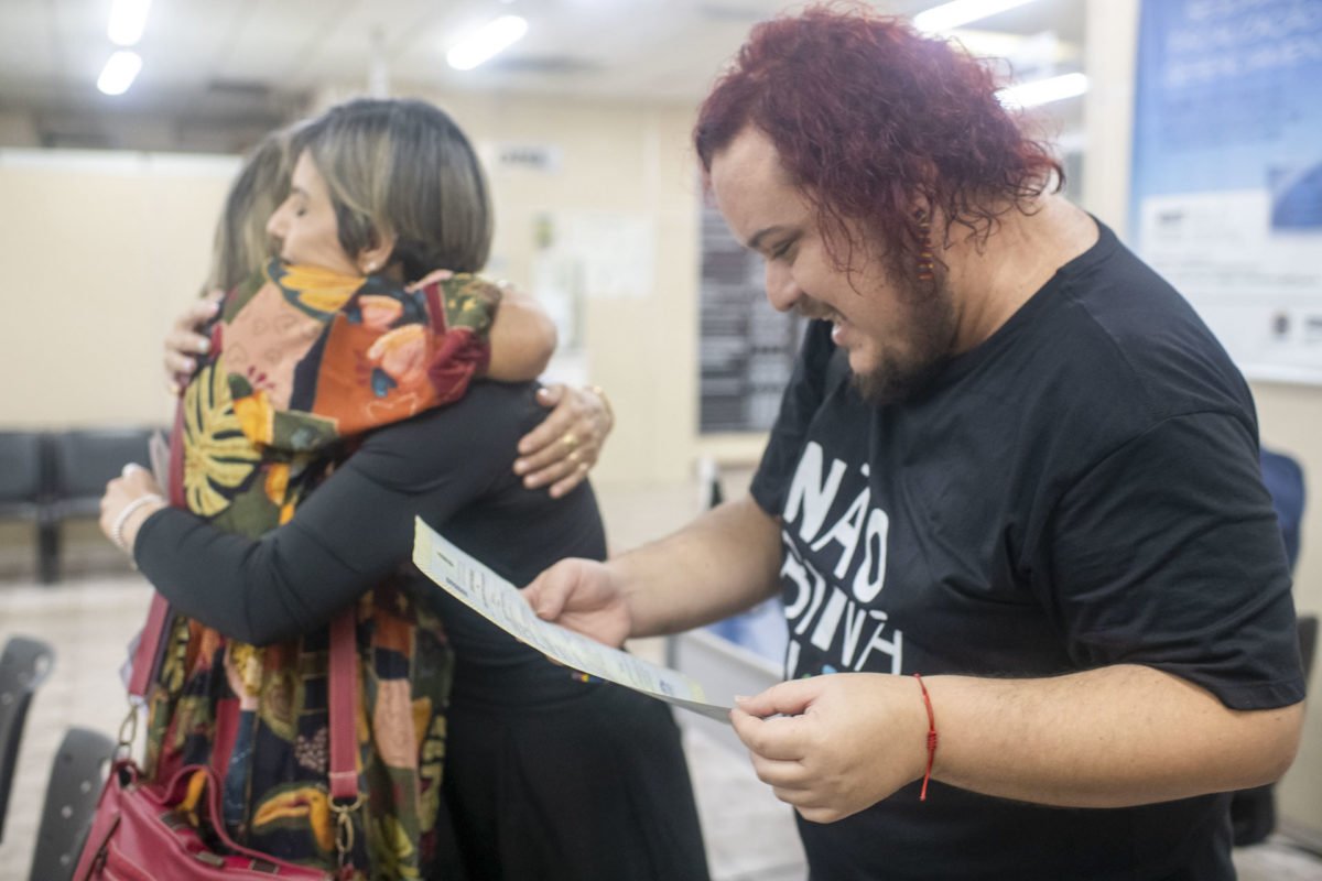 Homem branco de cabelo pintado de ruivo olhando para documento ao lado de mulher branca com cabelo curto e roupa preta que está abraçando outra mulher mais velha de roupa espantada