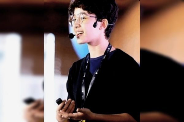 Foto colorida de jovem com óculos e microfone apresentando uma palestra