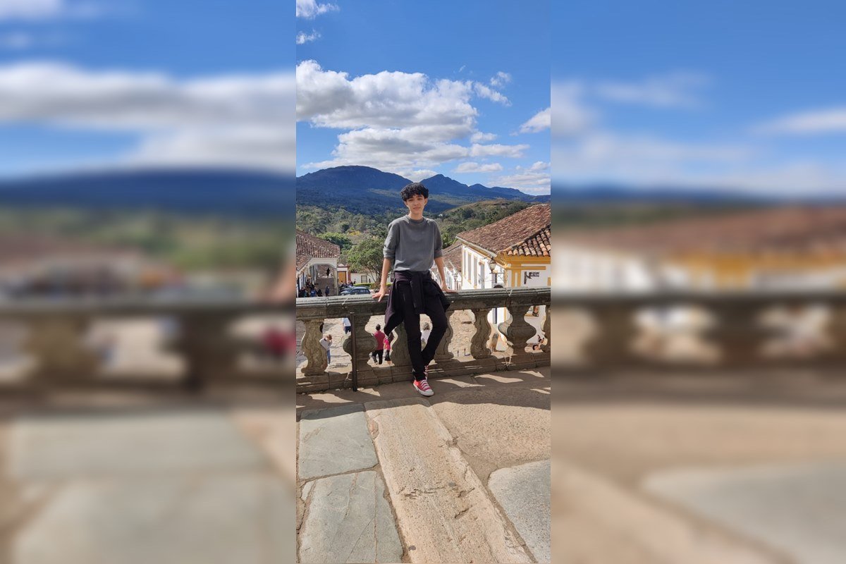 Foto colorida de jovem em frente a uma ponte com paisagem colonial ao fundo