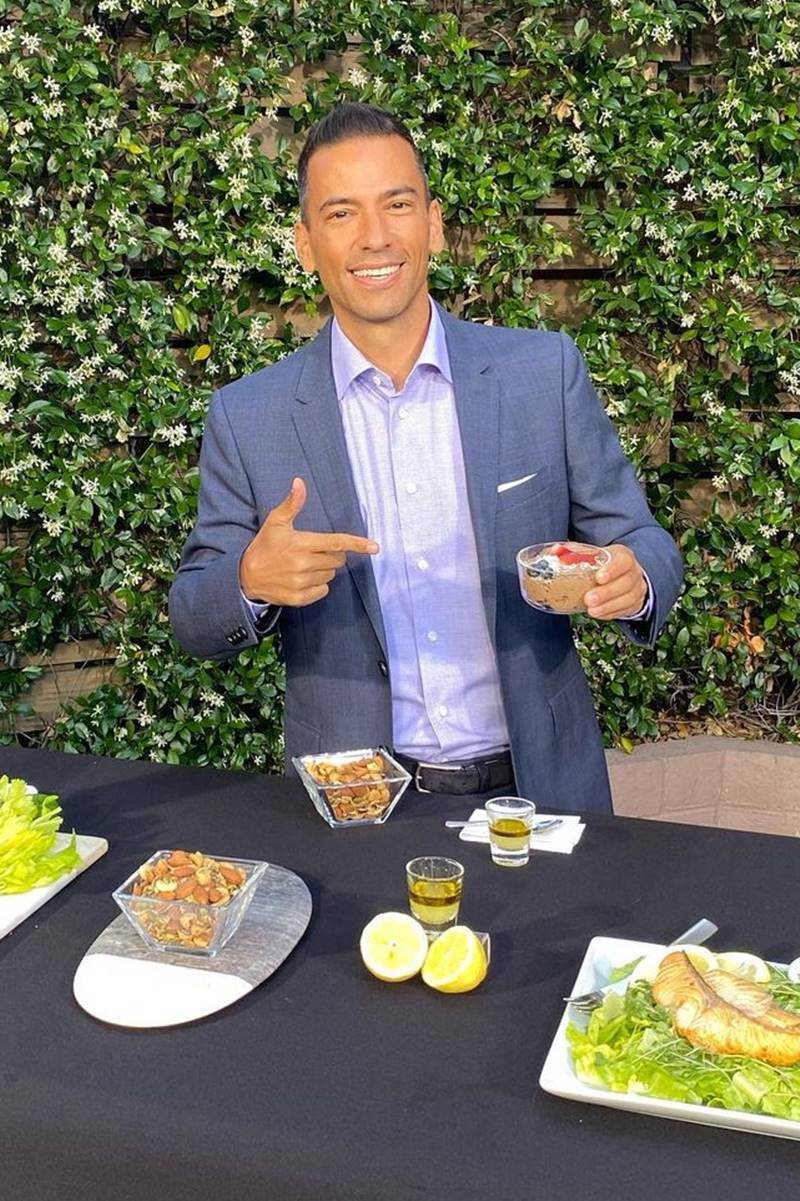 Foto de homem com blazer e blusa azul apontando para uma tigela com alimentos. Ele está atrás de uma mesa com alimentos - Metrópoles