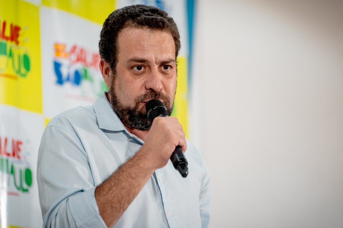 Imagem colorida mostra Guilherme Boulos falando ao microfone. Ele é um homem branco, de cabelo e barba castanhos, e veste camisa branca
