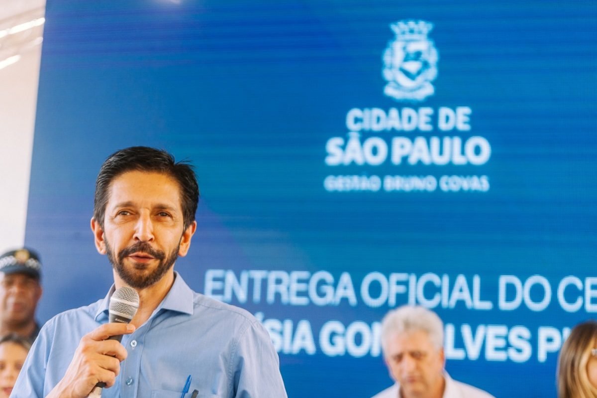 Imagem colorida mostra Ricardo Nunes, homem branco, de cabelo e barba pretos, em imagem do peito para cima, falando ao microfone na frente de um banner da Prefeitura - Metrópoles