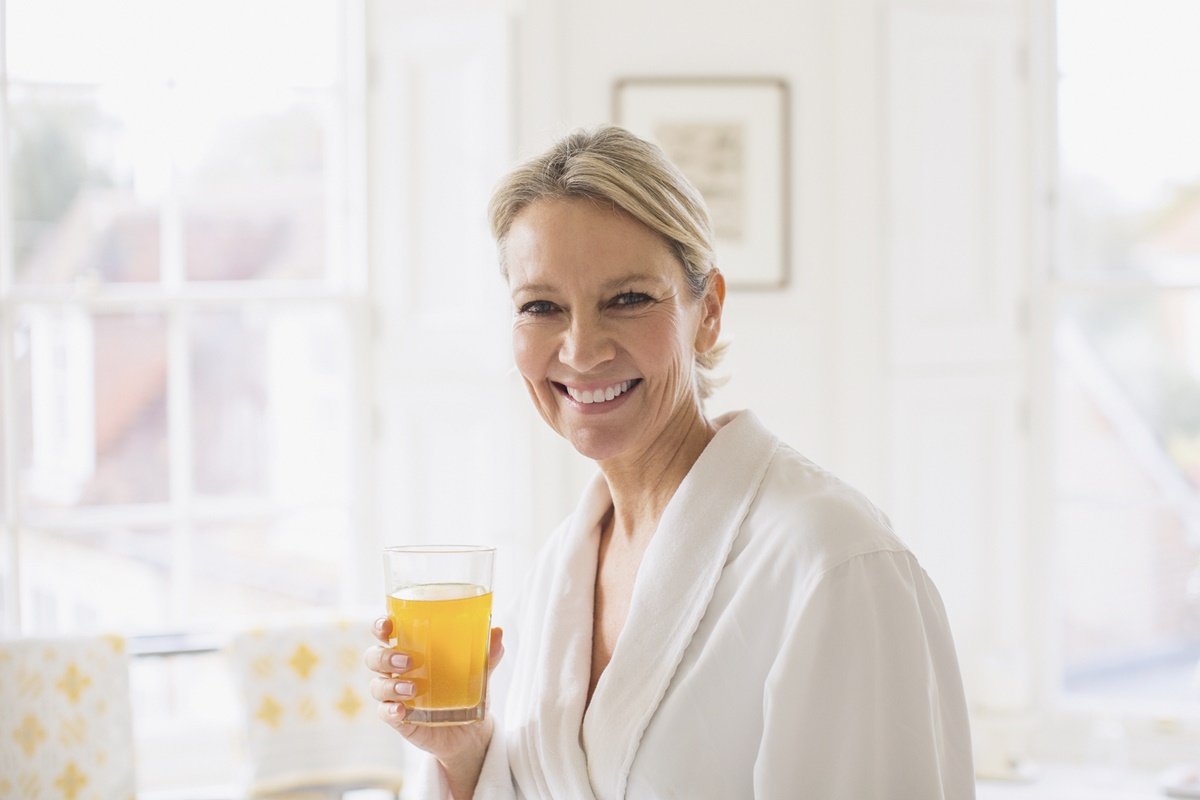 Foto de mulher idosa, vestida com roupão branco. Ela está sorrindo e segurando um copo transparente com líquido amarelado dentro - Metrópoles