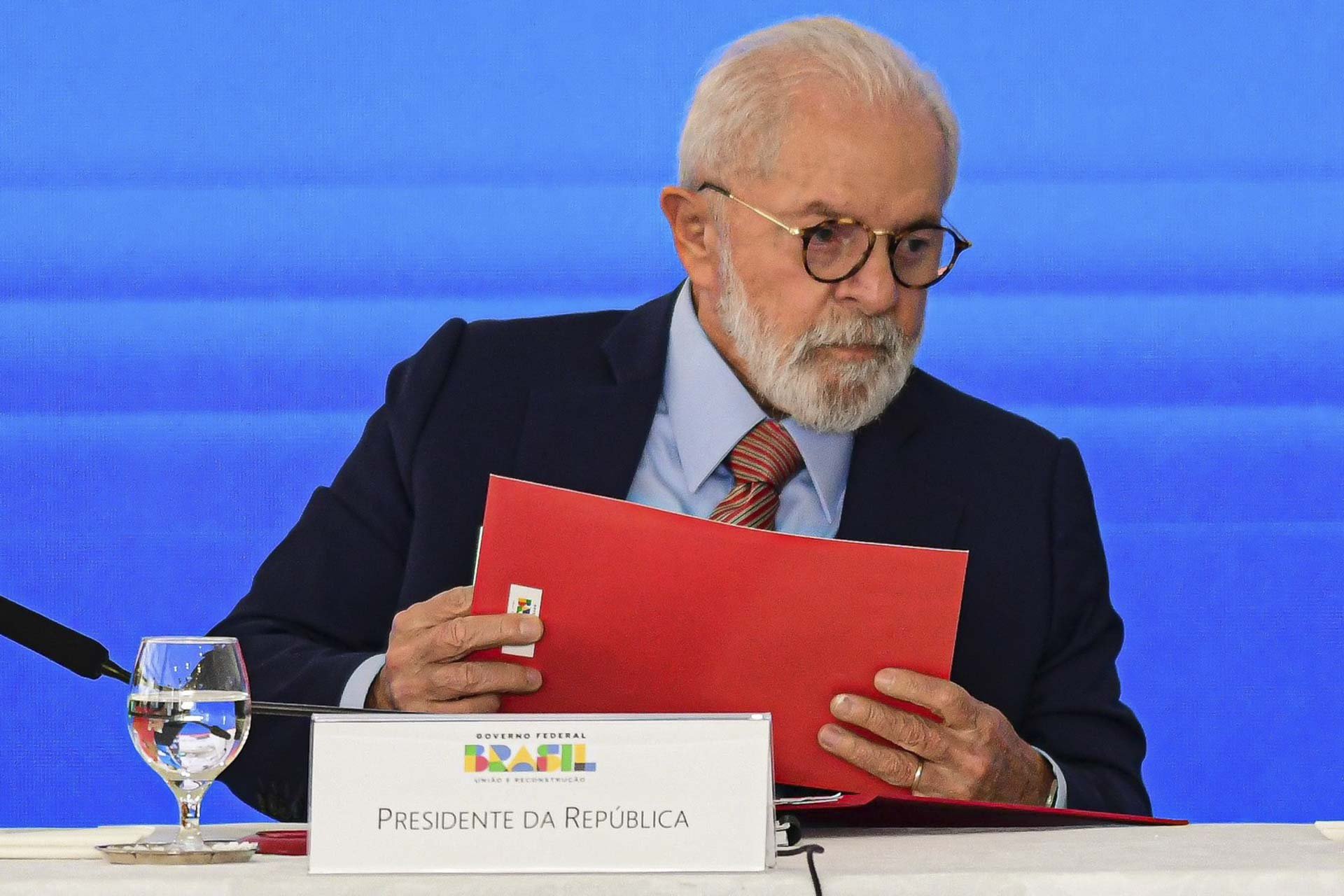 Imagem colorida do ex-presidente Lula usando óculos e segurando uma pasta vermelha - Metrópoles