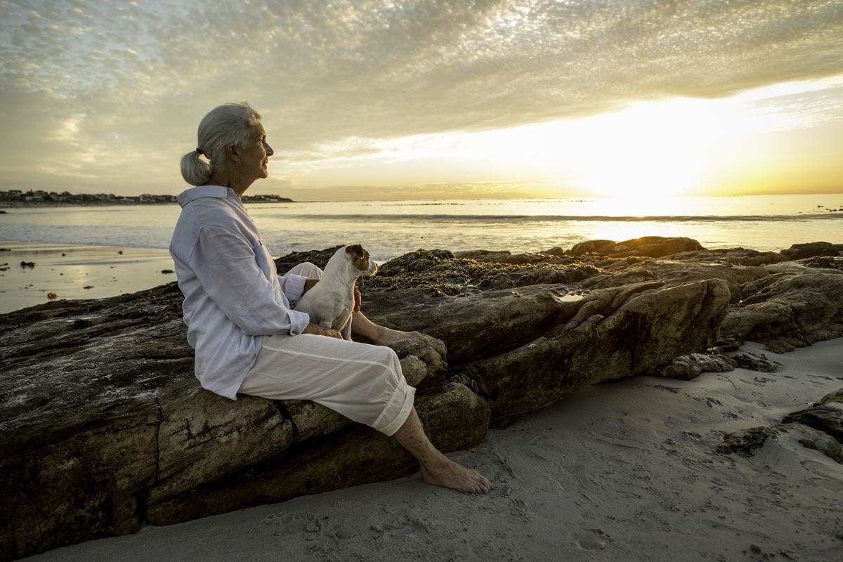 Foto colorida de mulher sentada em pedras, enquanto faz carinho de um cachorro e olha o pôr do sol - Metrópoles