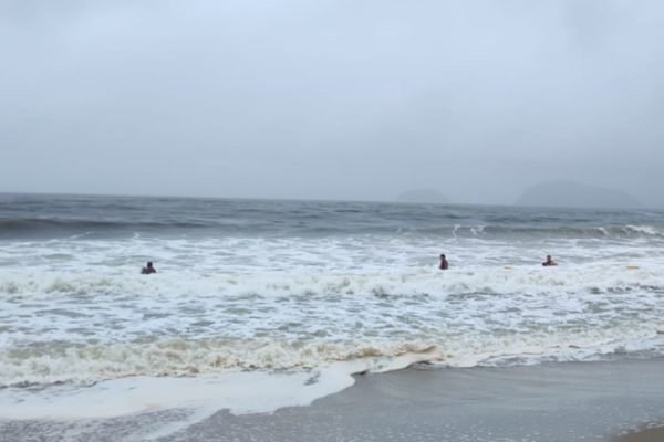 Foto colorida mostra mar cinzento e com ondas. Há três bombeiros na água buscando o corpo de duas garotas que desapareceram após serem levadas pela correnteza - Metrópoles