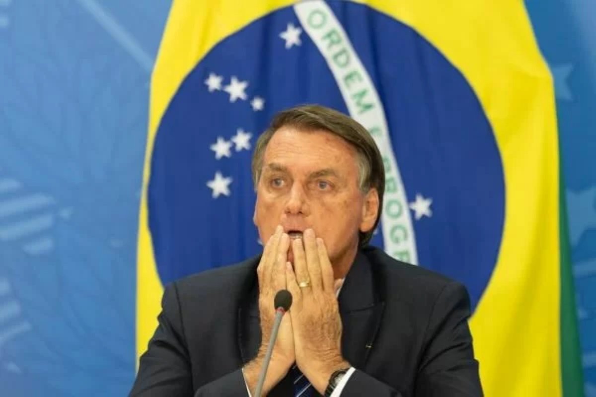 Foto colorida de Bolsonaro com mãos nas cabeça e fundo da bandeira do Brasil