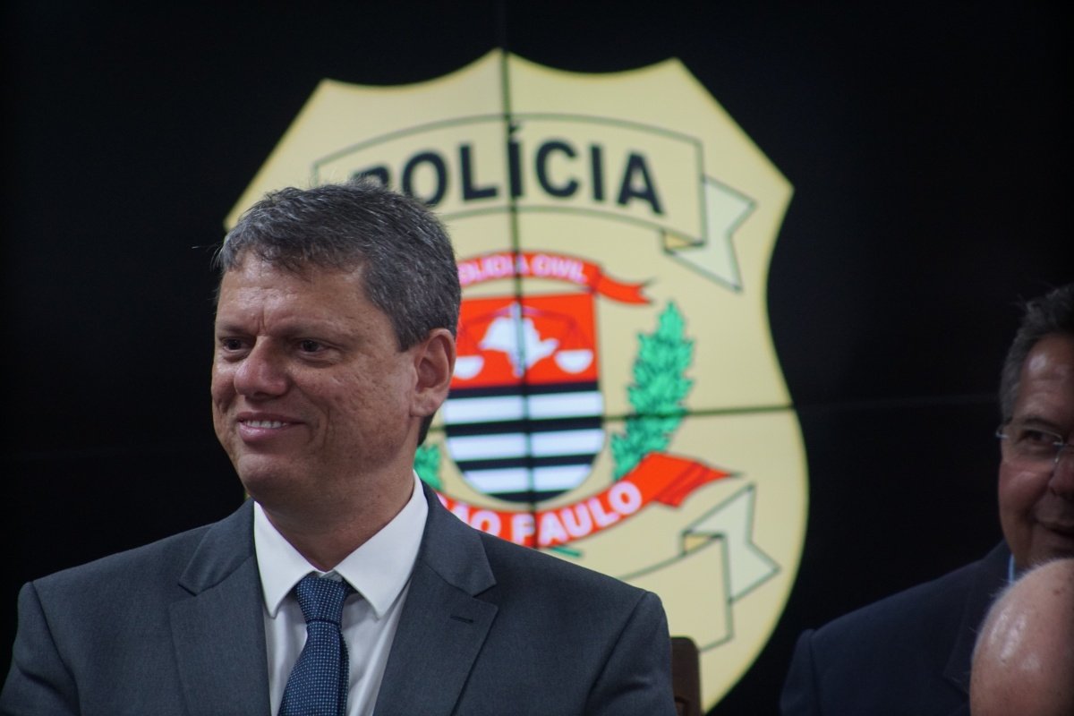 Imagem colorida mostra Tarcísio de Freitas, homem branco, grisalho, vestindo terno cinza, com um logotipo da Polícia Civil projetado em uma tela atrás dele - Metrópoles