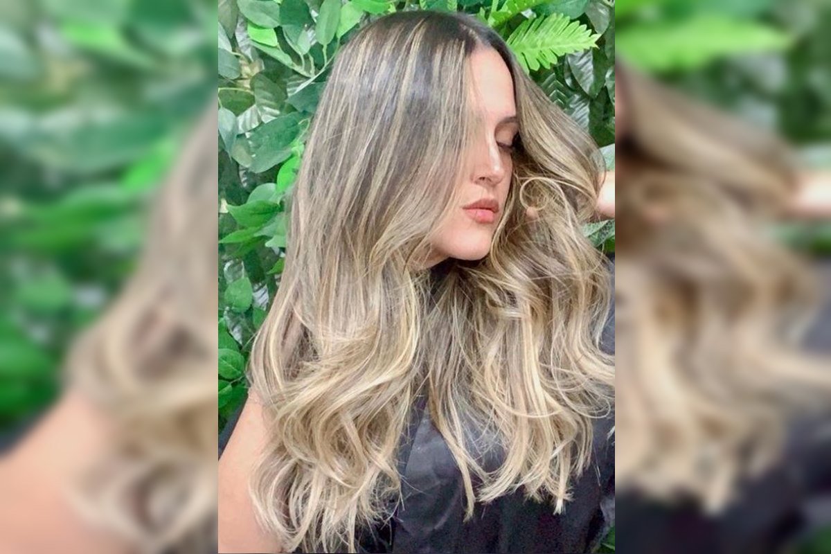 Foto colorida de mulher loira, com rosto lateralizado e plantas verdes ao fundo