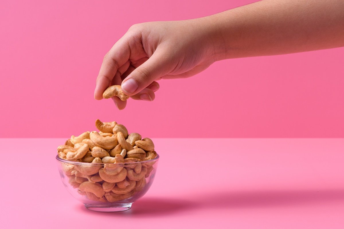 Foto colorida de mão pegando castanha-de-caju dentro de uma tigela de vidro. O fundo da fotografia é cor-de-rosa - Metrópoles