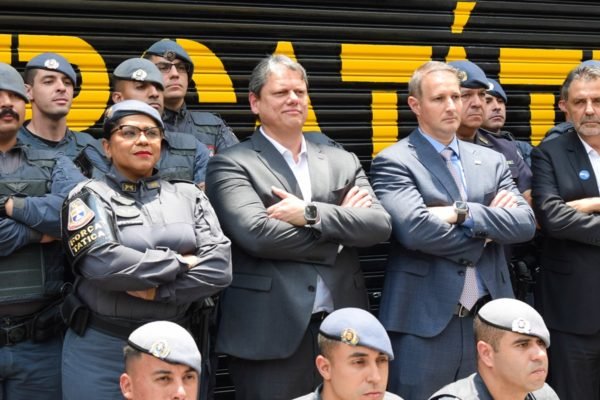 Imagem colorida mostra o governador Tarcísio de Freitas, de braços cruzados, enfileirado ao lado de PMs na mesma posição, na frente de um quartel da corporação - Metrópoles