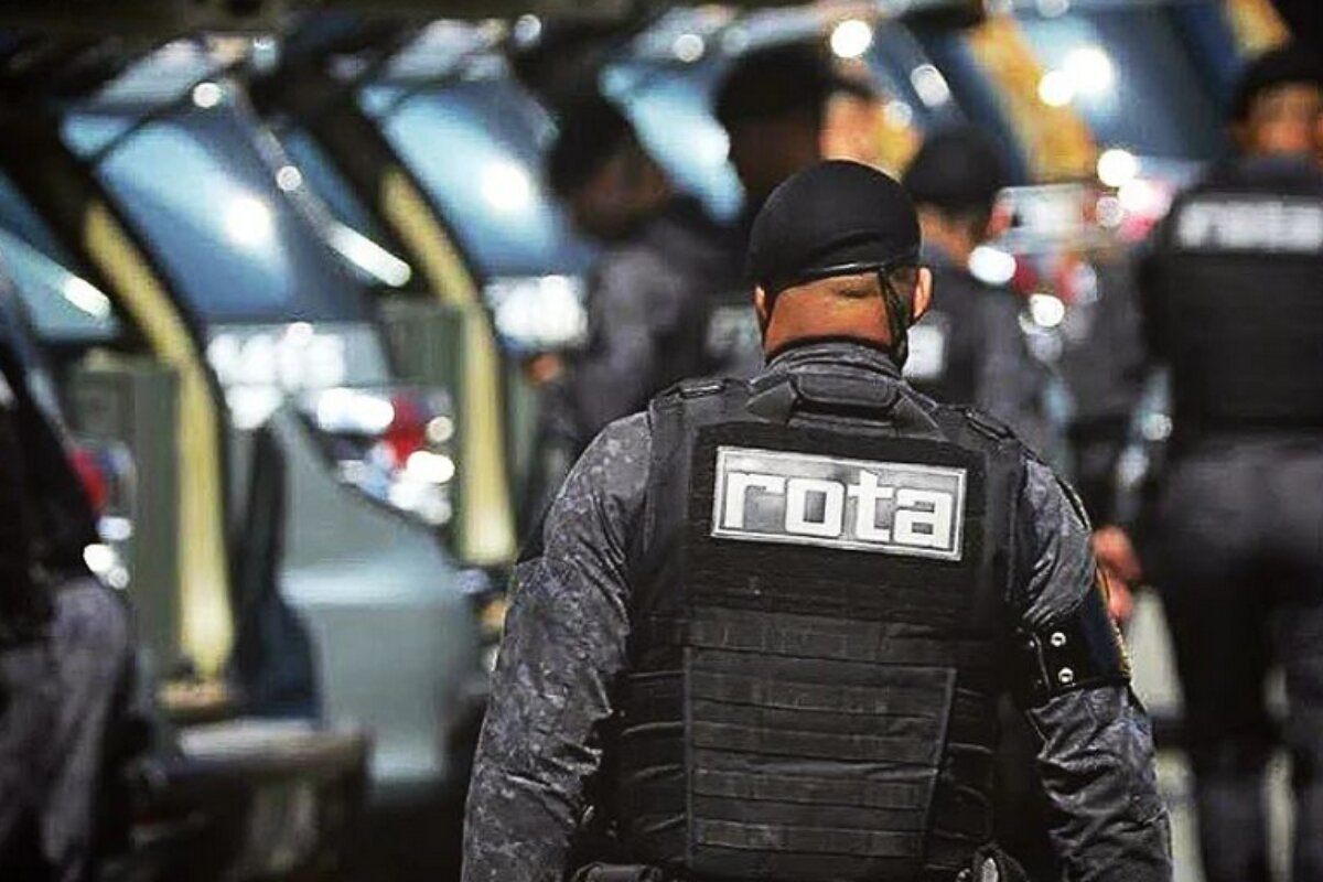 Em foto colorida policial de costas, na qual aparece a palavra Rota sobre o colete, com viaturas de fundo durante operação de combate à violência - Metrópoles