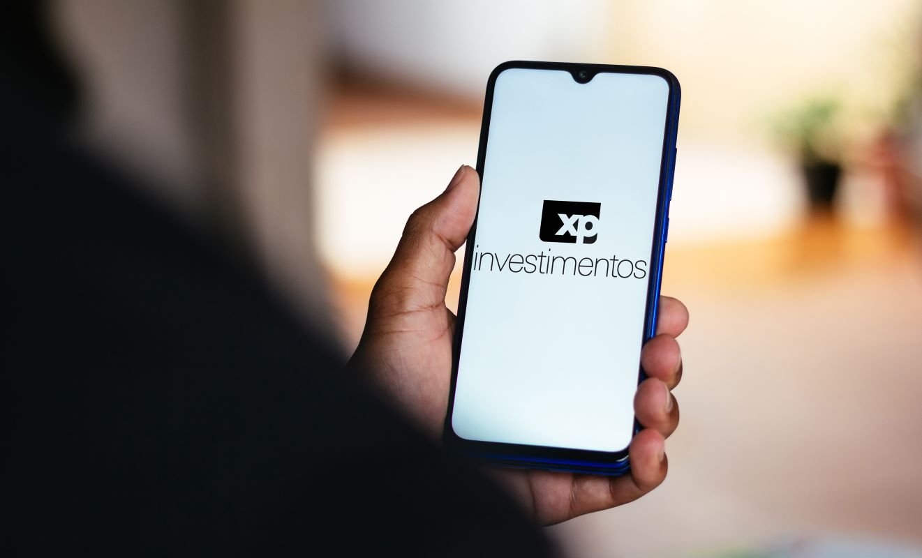 Imagem colorida de uma mão segurança um celular com logotipo da XP Investimentos na tela - Metrópoles