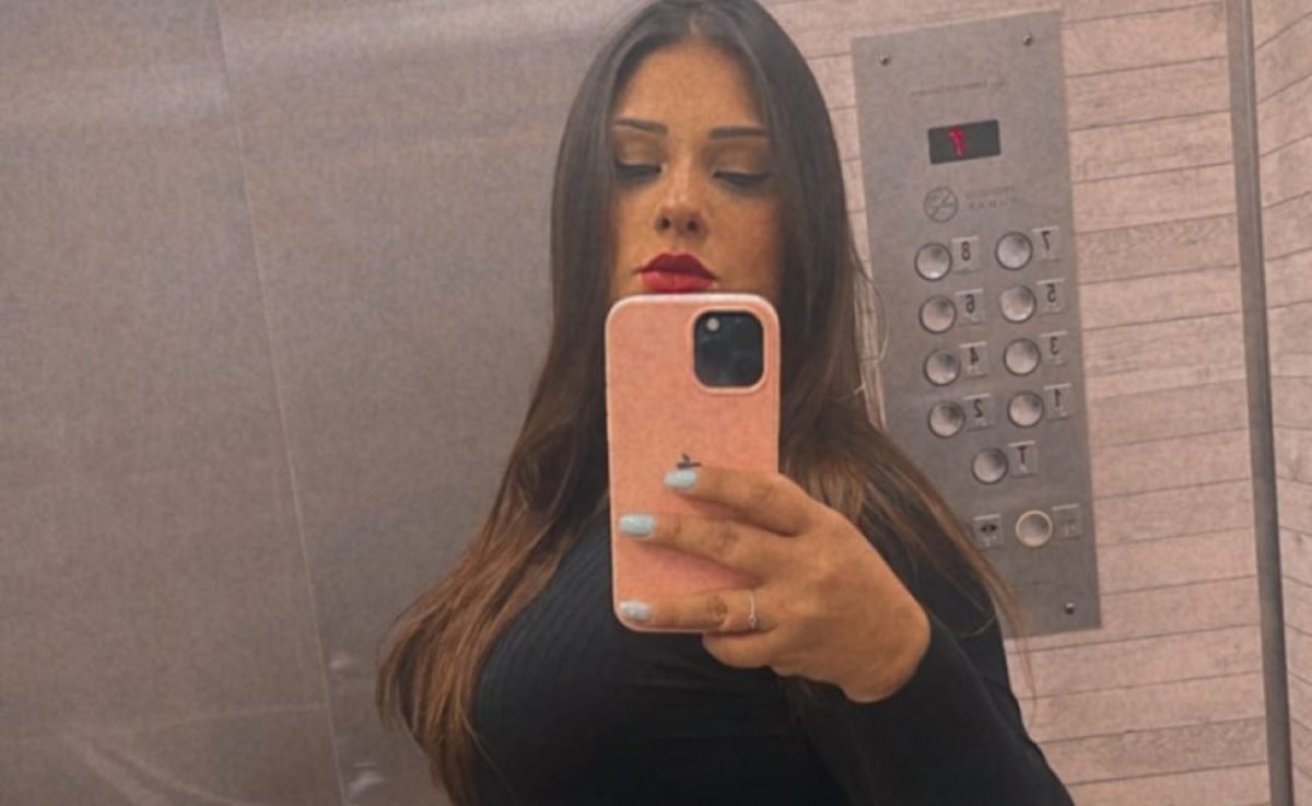 Mulher branca com cabelos escuros, batom vermelho e blusa preta segura celular em frente ao rosto