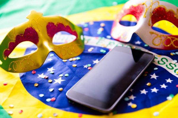 Na foto, celular sobre bandeira do brasil junto de máscaras de carnaval