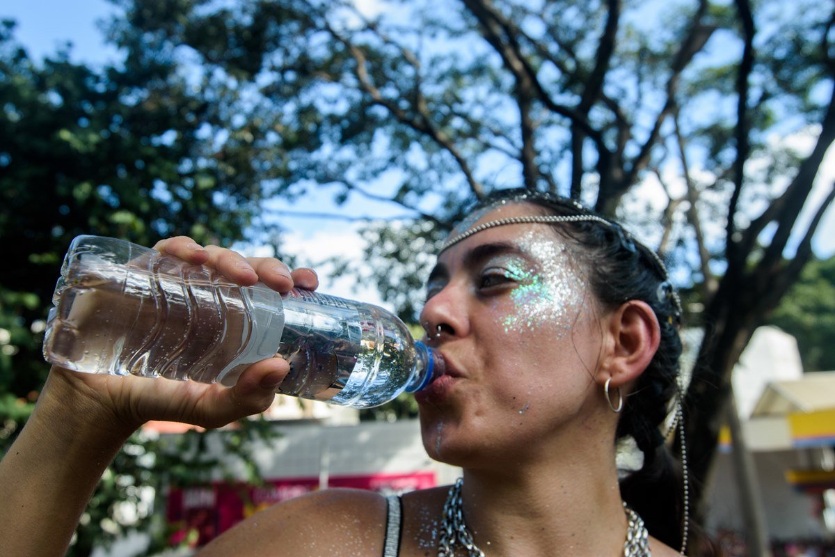 Fotografia mostra uma mulher com glitter no rosto bebendo água em uma garrafa transparente - Metrópoles