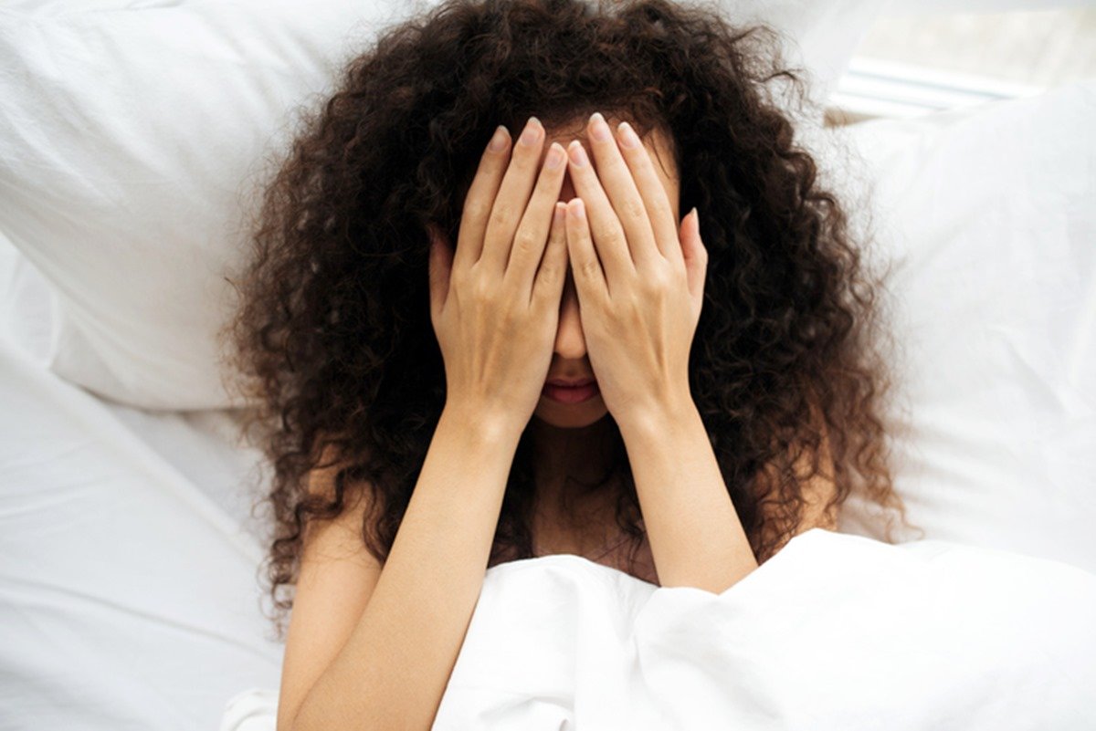 Mulher de cabelos cacheados com mão no rosto deitada em cama - Metrópoles