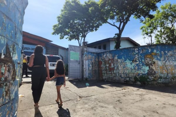 imagem colorida mostra mulher de mãos dadas com menina, estudante de uma escola. As duas estão de costas, andando em direção a uma escola - metrópoles