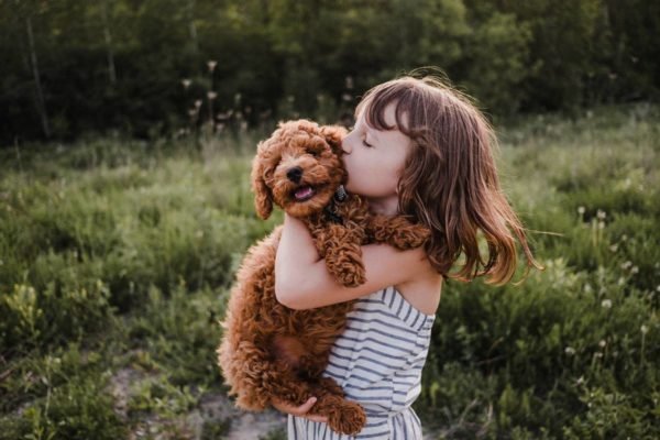 Foto colorida de uma menina abraçada com um cachorro marrom, o beijando no rosto - Metrópoles