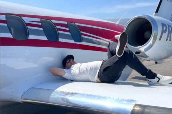 Homem de boné, óculos, calça preta e camiseta branca deitado sobre asa de avião de pequeno porte - Metrópoles