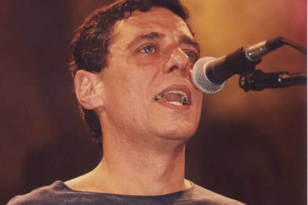 Foto colorida de Chico Buarque cantando em um microfone. Ele usa blusa marrom - Metrópoles