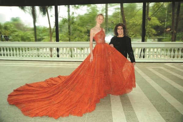 Modelo usa vestido laranjado logo com cauda e posa ao lado da estilista Martha Medeiros - Metrópoles