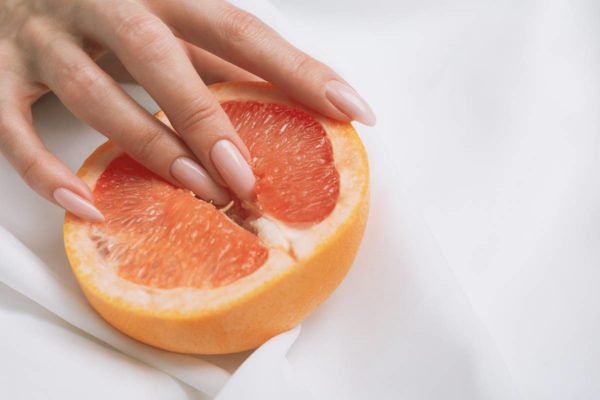 Foto colorida em close de dedos femininos com unhas grandes tocando uma grapefruit cortada no meio em um fundo branco - Metrópoles