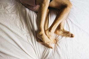 Foto colorida em close de quatro pernas de um casal deitado na cama com um lençol branco - Metrópoles