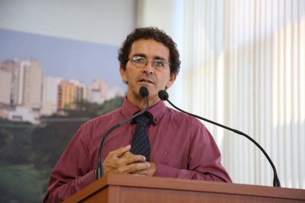 Homem pardo de óculos veste camisa de cor vinho e gravata preta enquanto fala em tribuna