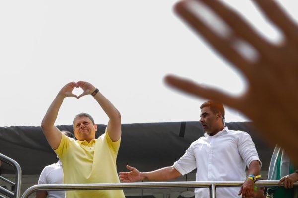 Imagem colorida de homem branco vestindo camiseta amarrela faz símbolo de coração com as mãos