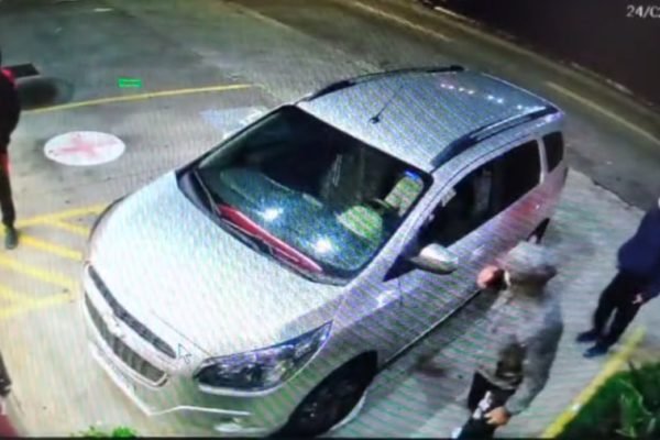 imagem colorida mostra cena filmada por câmera de segurança. três homens com moletons e máscaras estão ao redor de um carro de cor clara no estacionamento de uma farmácia - metrópoles