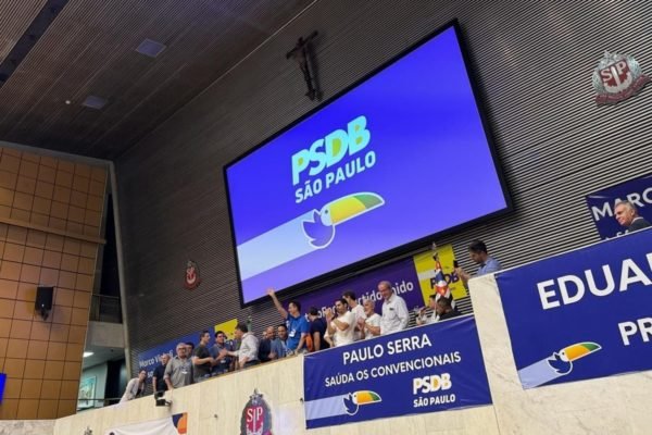 Imagem colorida mostra o plenário da Alesp com pessoas sorridentes e em pé na mesa diretora e uma série de faixas do PSDB