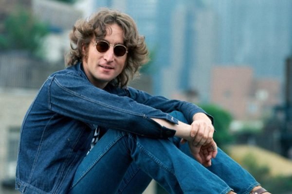 Foto colorida de John Lennon. Ele é um homem branco, usa jaqueta e calça jeans, óculos escuros e tem cabelo longo e loiro - Metrópoles