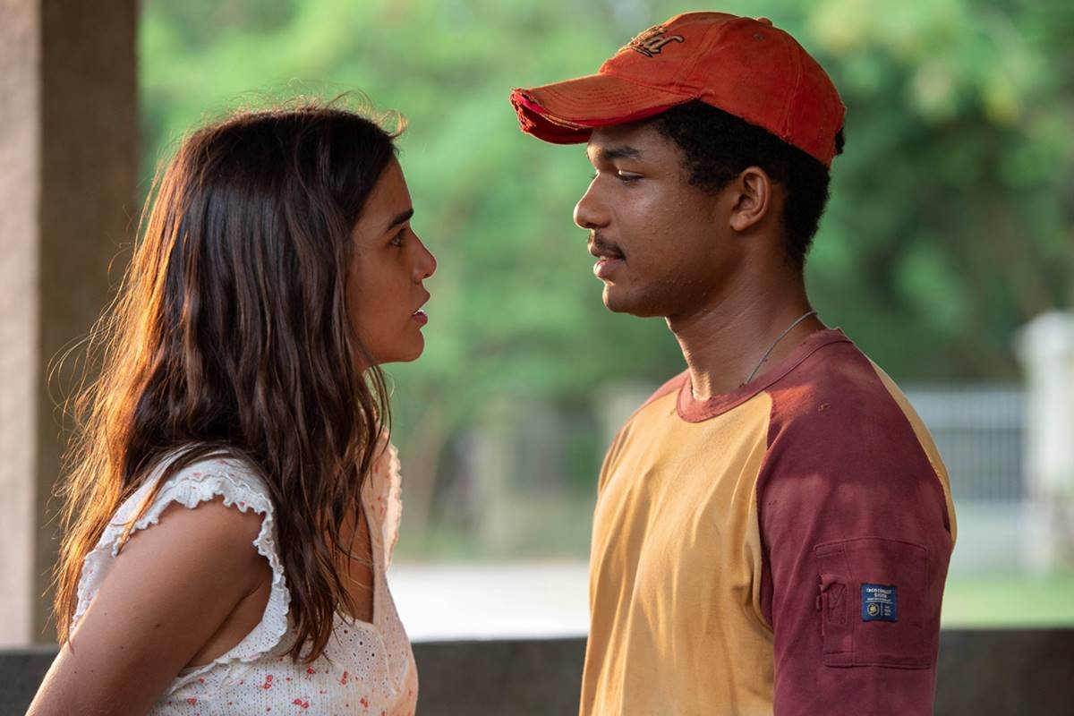 foto colorida de mulher de blusa branca e cabelos castanhos conversando com homem negro de camiseta amarela e vermelha e boné - metrópoles