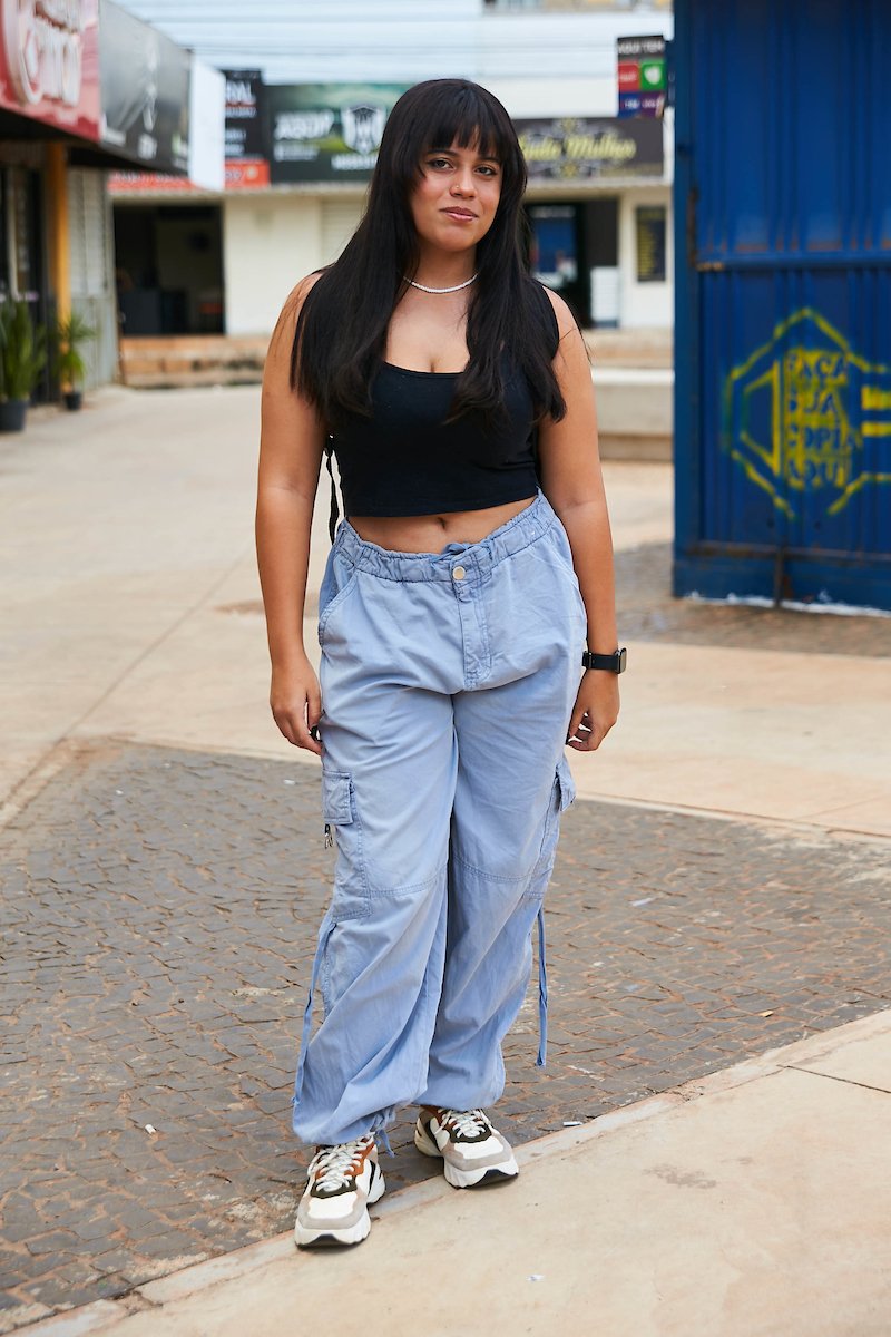 Garota de cabelo preto longo com franja posa para foto. Ela veste um top preto combinado com uma calça na cor cinza junto à seu tênis esportivo branco com detalhes em preto, cinza e marrom.