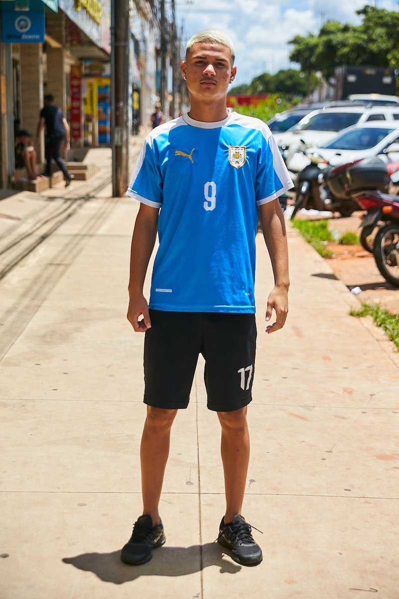 Jovem rapaz posa para foto. Olhando para a câmera, o garoto veste um conjunto de futebol de camisa azul com detalhes brancos e bermuda preta.