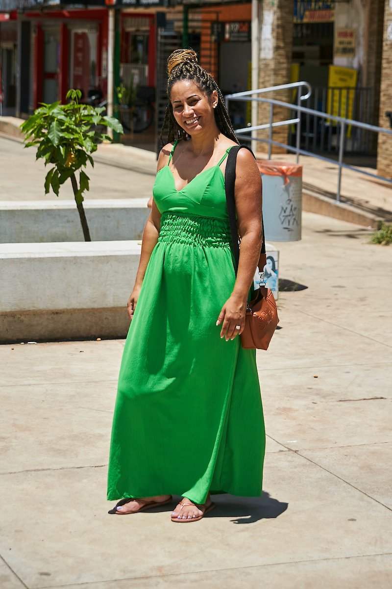 Mulher posa para foto com vestido verde. Em seu ombro esquerdo ela apoia sua bolsa de alça preta com tecido marrom.