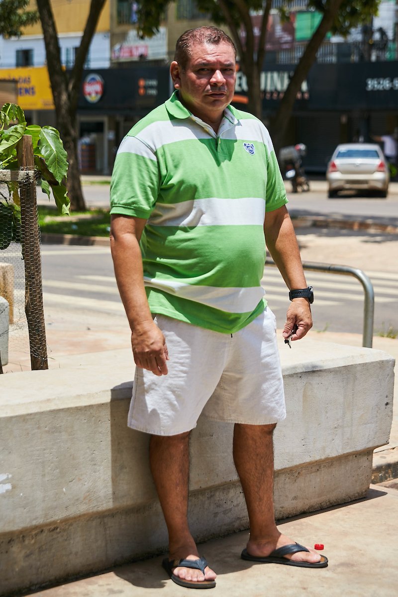 Homem posa para foto. Vestindo uma camisa polo verde com listras brancas e bordado azul, ele combina sua bermuda branca junto a seu chinelo preto. Em seu pulso esquerdo ele usa um relógio preto enquanto segura uma chave nas mãos.