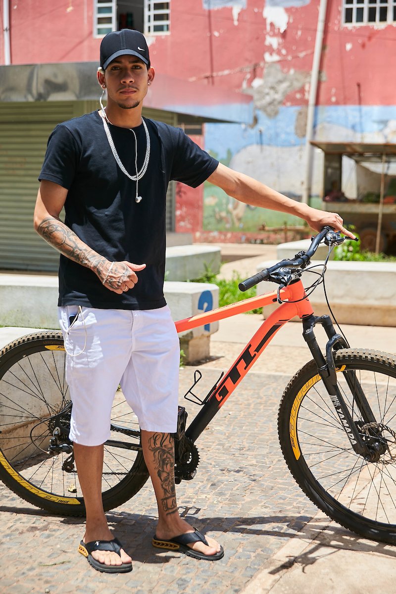 Rapaz posa para foto. De boné preto, ele veste uma camisa também preta combinada com as correntes em prata e uma bermuda branca. Nos pés, ele calça uma sandália preta com detalhes dourados que combinam com a bicicleta apoiada em sua mão esquerda.