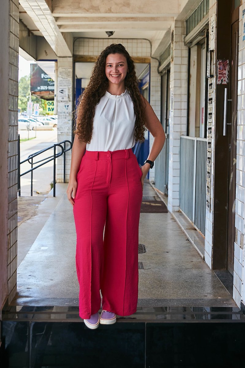 Mulher jovem combina camisa branca com calça rosa. No pulso esquerdo ela porta seu relógio na cor preta. Calçando um par de tênis roxo ela combina o sapato com detalhe vermelho com a calça.