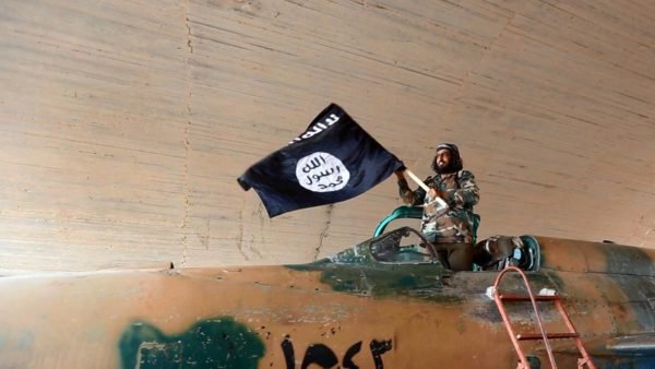 Imagem colorida mostra combatente do Estado Islâmico em um tanque segurando a bandeira do grupo - Metrópoles