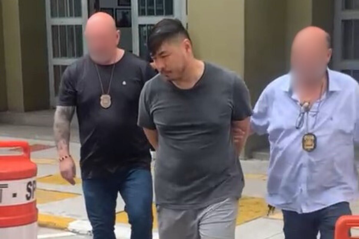 Em foto colorida homem japonês com sobrepeso no centro é conduzido, com as mãos algemadas para trás, por dois policiais civis - Metrópoles