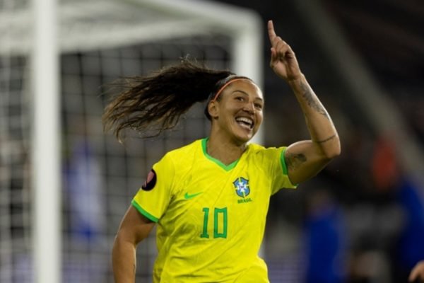 Foto mostra a atacante brasileira Bia Zaneratto com a camiseta amarela da seleção feminina, apontando para cima com um gol ao fundo - Metrópoles