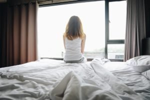Foto colorida de uma mulher de costas sentada na beirada da cama olhando pela janela - Metrópoles