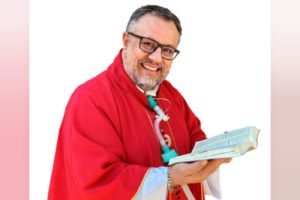 Imagem de fundo borrado mostra padre com vestimenta vermelha e com a bíblia na mão. Ele tem traqueostomia por sua doença rara - Metrópoles